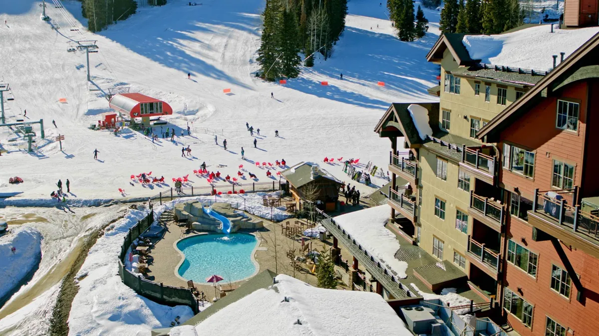 A vibrant winter scene showing a swimming pool adjacent to ski slopes and a resort building. Lounge chairs border the pool, while guests relax poolside, and the ski lift and snowy mountains are visible in the background. The atmosphere is lively and energetic, capturing guests enjoying both aquatic and winter activities.