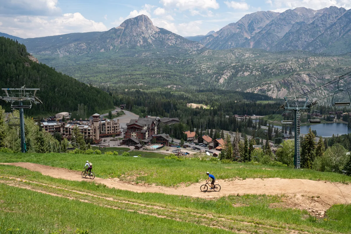 This vibrant exterior scene captures the mountains surrounding the resort, with visible ski lifts, bike trails, resort buildings, and a sparkling lake. The image conveys the adventure and natural beauty available right outside the property.