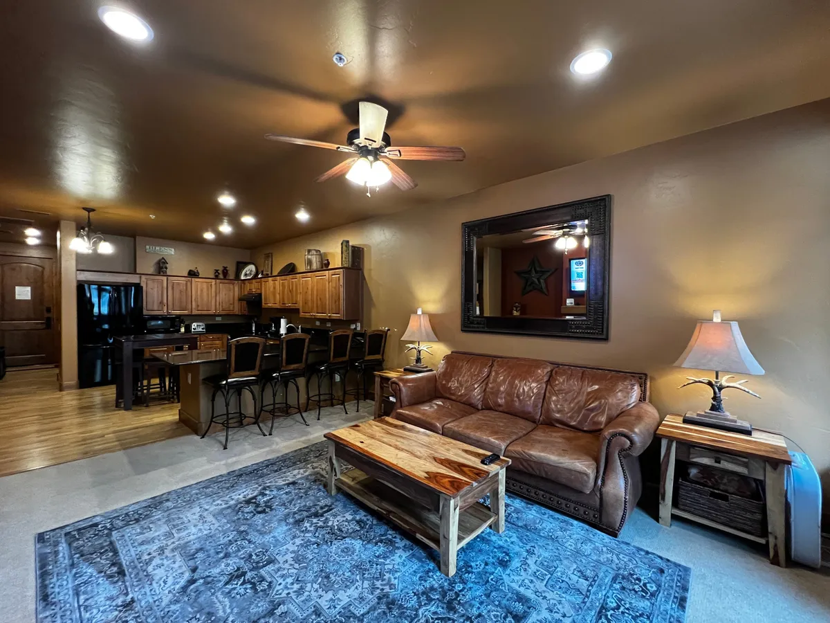 This open-plan interior features a comfortable leather sofa, a wooden coffee table, and a kitchen area with a dining bar. The view allows a glimpse of entertaining spaces that lead directly into the kitchen, offering a sense of flow for gatherings and celebrations.