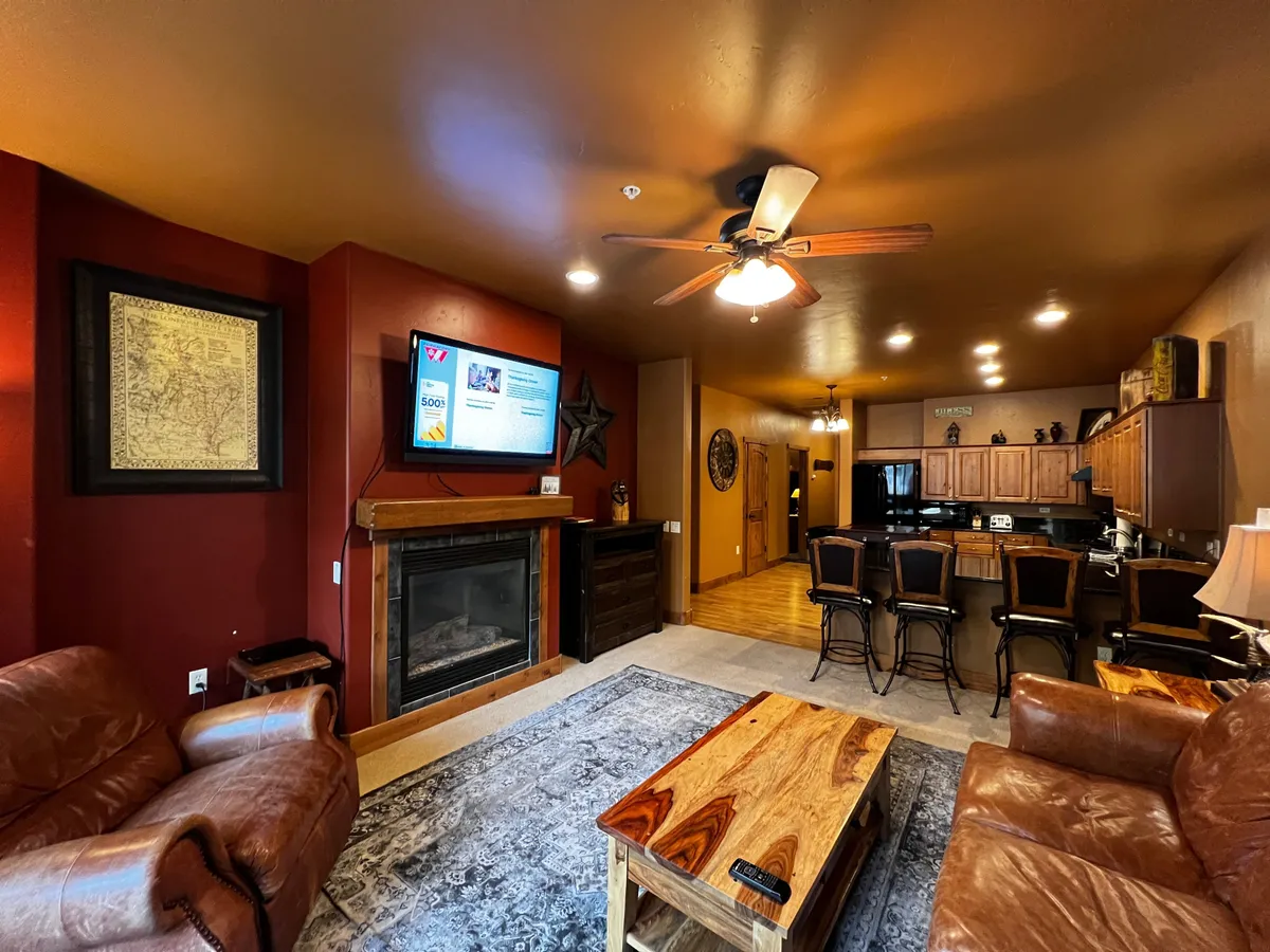 The image showcases a cozy living room interior featuring a crackling fireplace, comfortable leather sofas arranged for conversation, a stylish wooden coffee table, and a large television. The warm lighting and inviting setup suggest an ideal spot for relaxing, gathering with friends or family, and enjoying leisure activities like game nights or movie marathons.