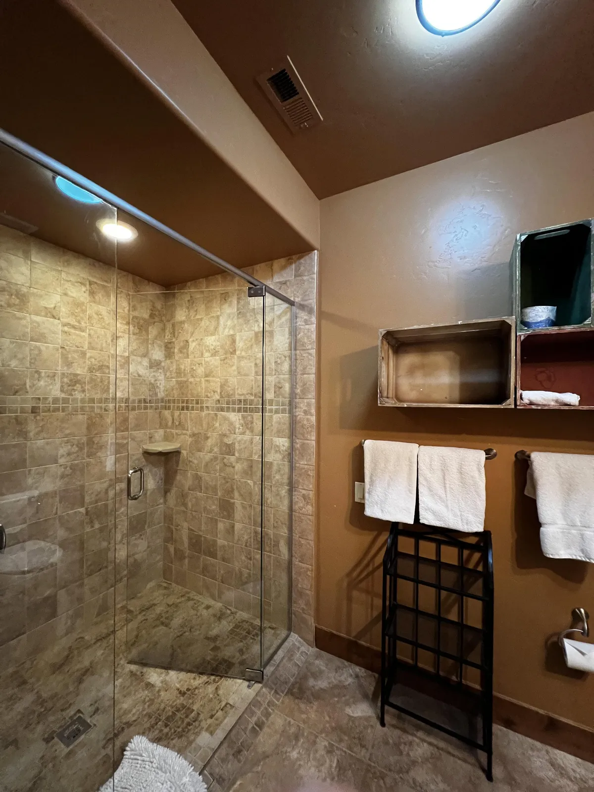 A modern bathroom featuring a glass shower enclosure, textured tile walls, neatly arranged towels, and open storage boxes. The space feels organized, tranquil, and spotlessly clean—ideal for relaxation and self-care.