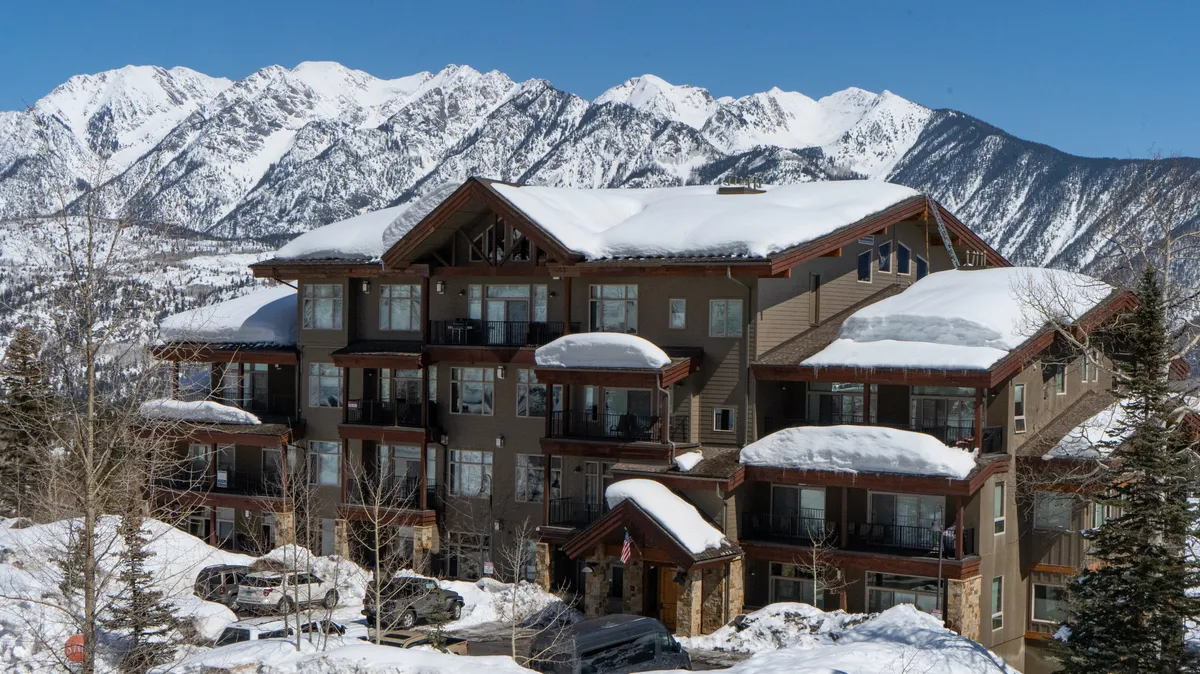 A cozy exterior view of the property with snow-covered roofs, balconies overlooking the mountains, and people enjoying the snowy landscape. Parked cars and mountain backdrops suggest easy access to winter sports and après-ski relaxation.