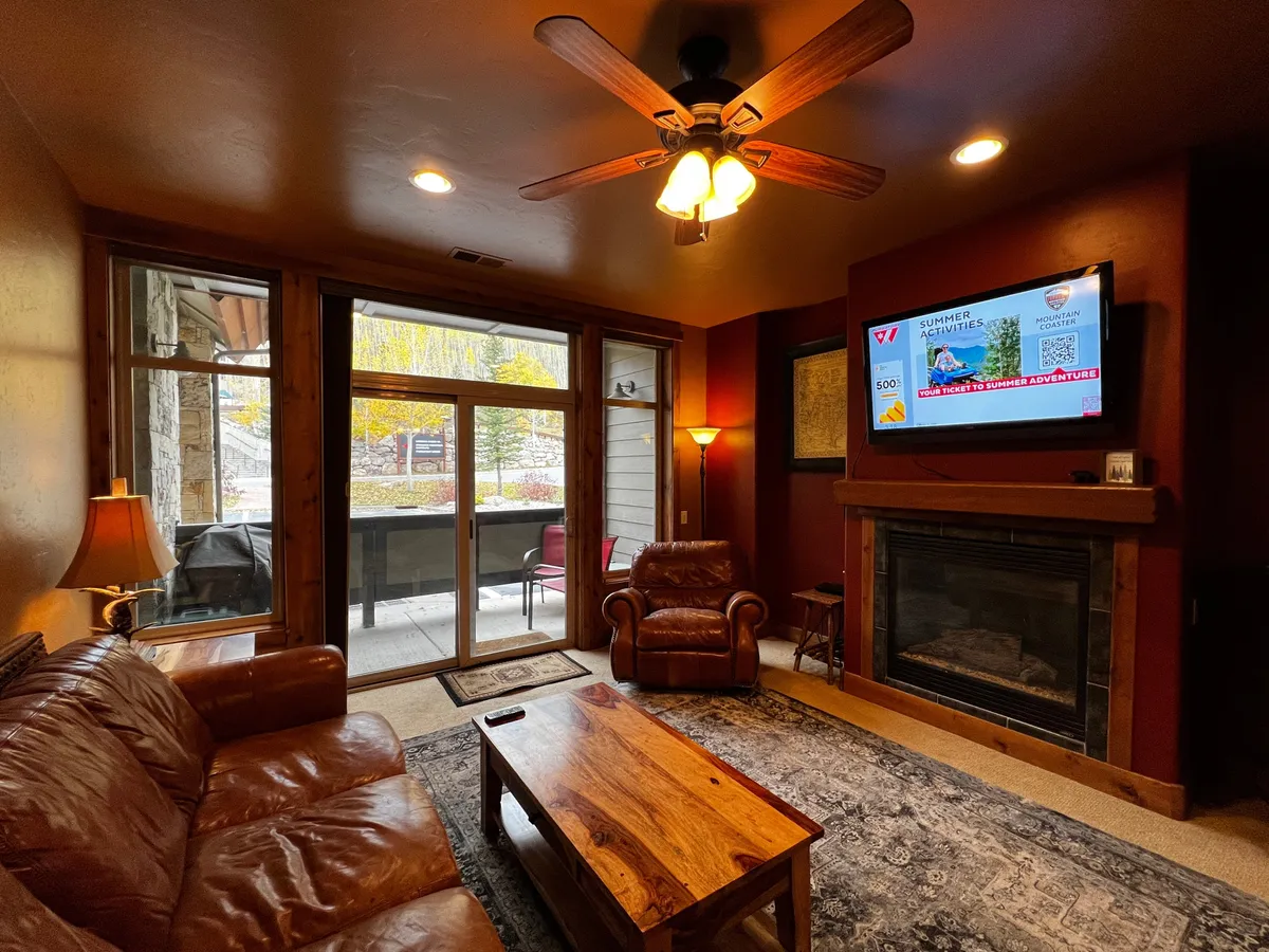 This inviting interior features a leather sofa, wood coffee table, a cozy fireplace, a large television, and expansive windows that let in plenty of natural light. The plush seating arrangement faces the entertainment center, while the view outside hints at mountain scenery.