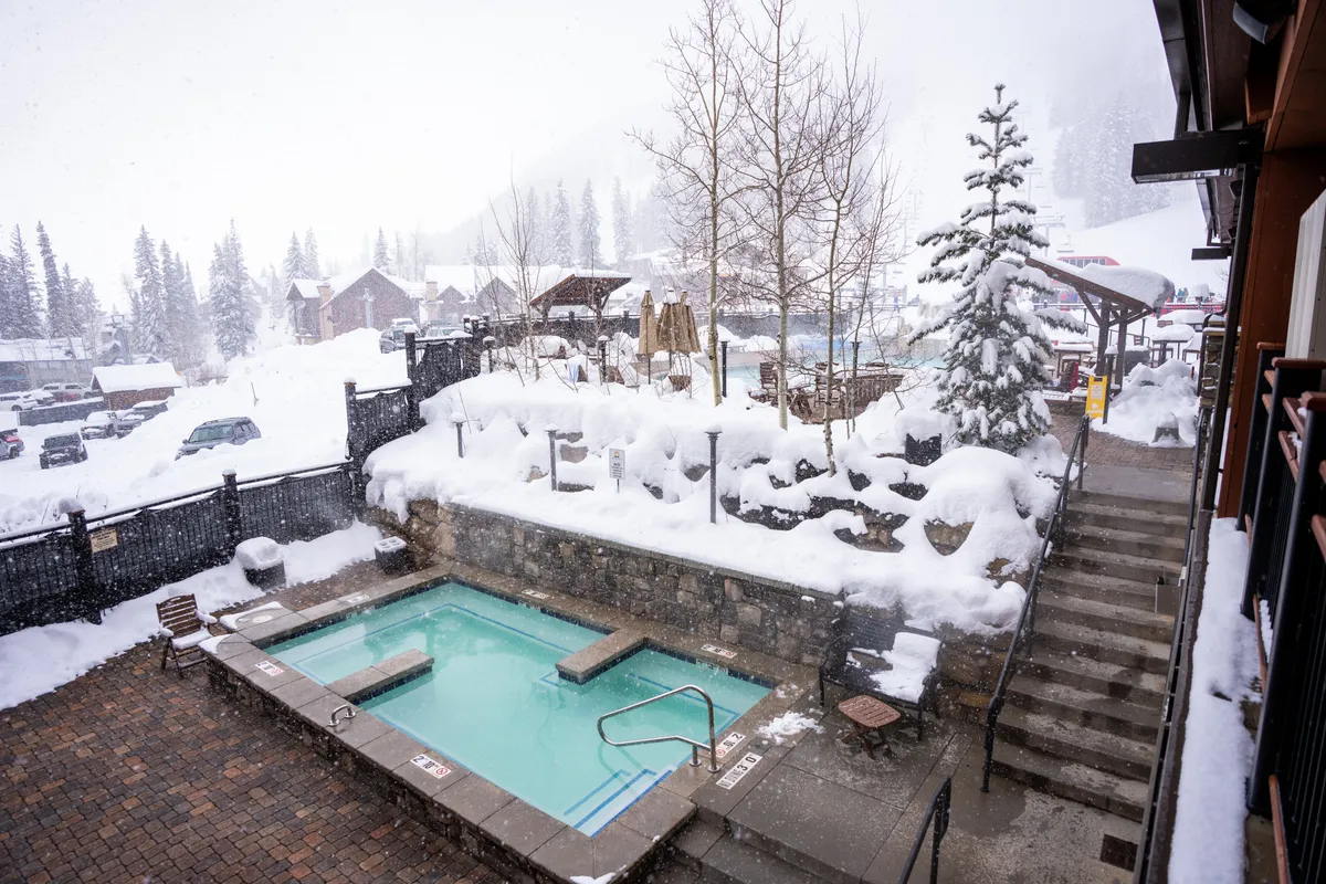 An inviting outdoor hot tub surrounded by snow-covered trees and a rustic stone wall, with a staircase leading to the tub area. Steam rises from the bubbling water as people enjoy the warmth amidst a tranquil, wintry landscape.